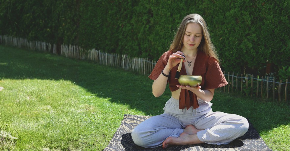 Sophie sitzt im Schneidersitz auf einer Decke im Garten und hält eine Klangschale