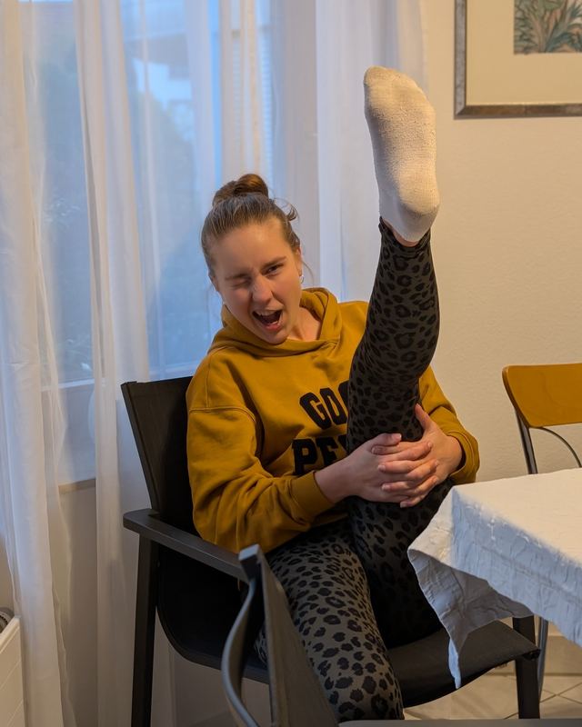 Sophie sitzt lachend auf einem Stuhl am Esstisch, hält ein Bein senkrecht nach oben gestreckt und trägt dazu den senffarbenen Hey-Honey-Hoodie