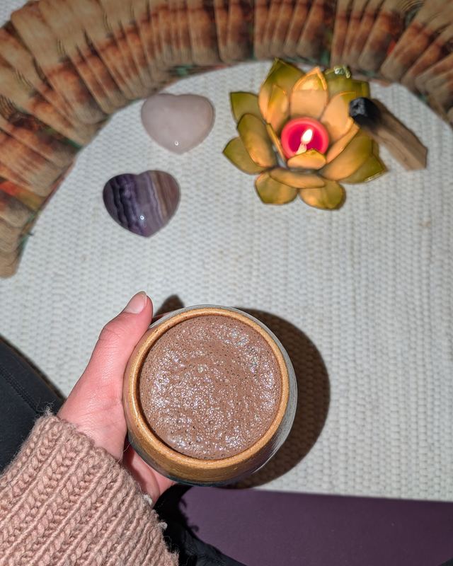 Hand hält einen Becher mit schaumigem Kakao, daneben auf heller Decke zwei Kristallherzen und eine brennende Lotuskerze