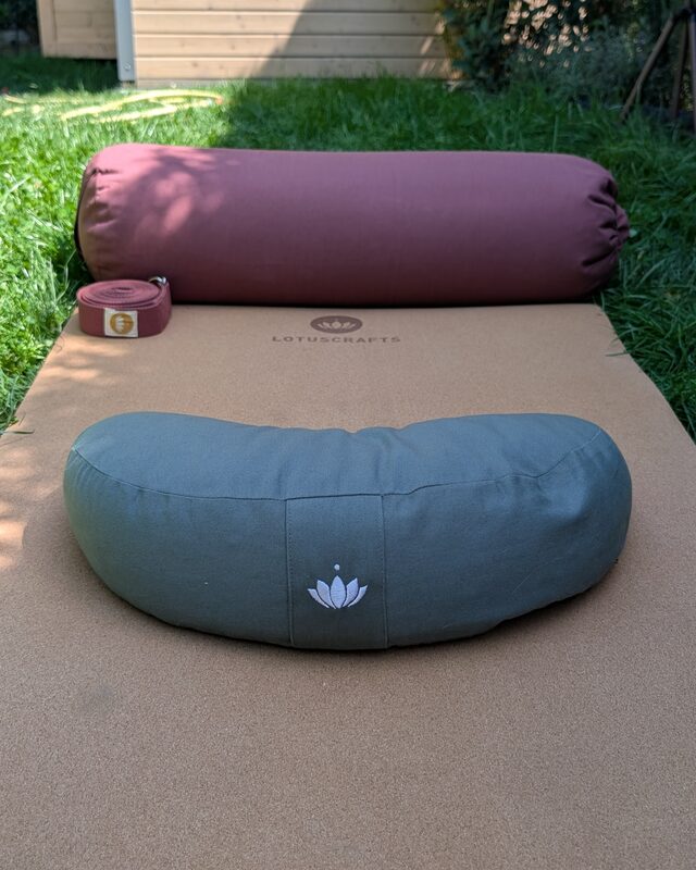 Ein weinrotes Bolster, ein aufgerollter Yogagurt und ein graugrünes Halbmondkissen auf einer Korkmatte im Garten