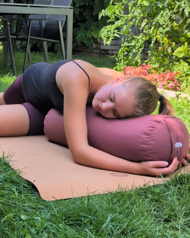 Sophie liegt mit geschlossenen Augen vornüber auf dem Bolster, auf einer Korkmatte im Gras